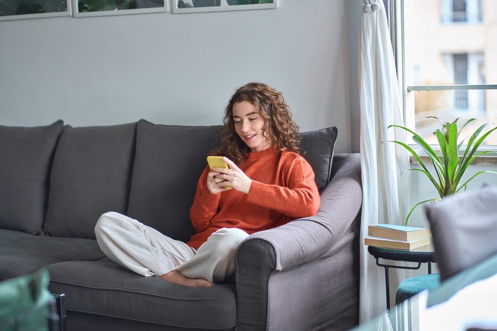 Smiling young woman sitting on sofa using mobile phone at home.